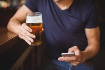 Sección media del hombre usando el teléfono móvil mientras toma un vaso de cerveza en el bar - foto de stock