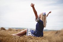 Беззаботная блондинка сидит в поле с поднятыми руками — стоковое фото