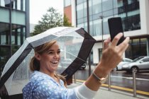 Belle femme tenant parapluie tout en prenant selfie pendant la saison des pluies — Photo de stock