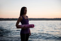 Beautiful woman standing with yoga mat on beach at dusk — Stock Photo