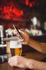 Uomo con bicchiere di birra utilizzando il telefono cellulare in bancone al bar — Foto stock