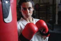 Portrait of woman in karate kimono and boxing gloves standing near punching bag in fitness studio — Stock Photo
