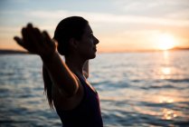 Vue latérale de la femme attrayante exécutant le yoga sur la plage pendant le coucher du soleil — Photo de stock