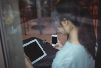Vista trasera de la mujer utilizando el teléfono móvil y la tableta digital en la cafetería - foto de stock