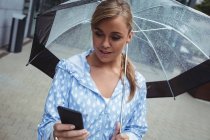 Beautiful woman holding umbrella while using smartphone during rainy season — Stock Photo