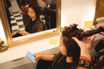 Femme utilisant un téléphone portable tout en se faisant lisser les cheveux au salon de coiffure — Photo de stock