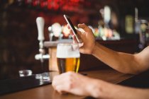 Homem com copo de cerveja usando telefone celular no balcão no bar — Fotografia de Stock