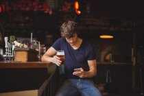 Hombre usando el teléfono móvil mientras toma un vaso de cerveza en el bar - foto de stock