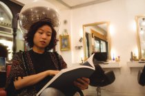 Elegante donna che legge una rivista mentre siede sotto un asciugacapelli al salone di parrucchiere — Foto stock