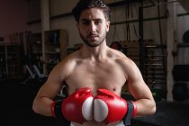 Porträt eines hemdslosen Boxers mit Boxhandschuhen, der im Fitnessstudio in die Kamera blickt — Stockfoto