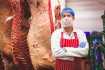 Retrato del carnicero de pie con los brazos cruzados en la sala de almacenamiento de carne en la carnicería - foto de stock