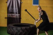 Vue latérale du boxeur thaïlandais frappant pneu avec marteau de traîneau dans la salle de fitness — Photo de stock