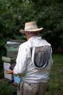 Rear view of beekeeper holding beehive in wooden frame at apiary garden — Stock Photo