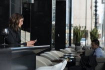 Femme d'affaires utilisant un téléphone portable au comptoir du bar tandis que l'homme d'affaires utilisant une tablette numérique sur le canapé de l'hôtel — Photo de stock