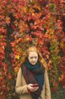 Redhead woman using mobile phone against plant creeper during autumn — Stock Photo