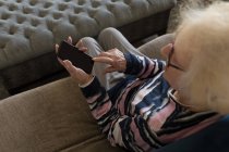 Femme âgée utilisant un téléphone portable sur le canapé dans le salon à la maison — Photo de stock