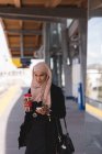 Хиджаб женщина с помощью мобильного телефона за чашкой кофе на платформе на железнодорожном вокзале — стоковое фото