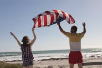 Vista trasera de pareja sosteniendo bandera americana en la playa - foto de stock