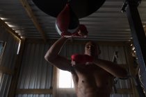 Visão de baixo ângulo do boxeador masculino praticando boxe com saco de perfuração no clube de boxe — Fotografia de Stock