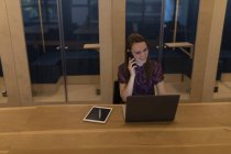 Деловая женщина разговаривает по мобильному телефону во время использования ноутбука на рабочем столе в офисе — стоковое фото