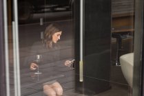 Mujer de negocios tomando vino tinto mientras usa el teléfono móvil en el hotel - foto de stock