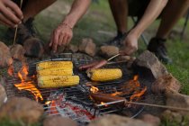 Primer plano de hombres asando salchichas y maíz en fogata en el camping - foto de stock