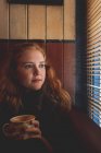 Продумана руда жінка дивиться крізь вікно сліпим у кафе — стокове фото