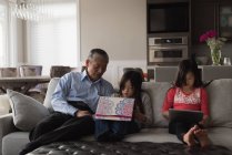 Abuelo y nietas usando tableta digital en el sofá en la sala de estar en casa - foto de stock