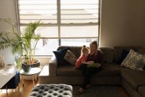 Madre e hija usando tableta digital en el sofá en la sala de estar en casa - foto de stock