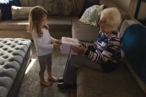 Nieta dando regalo a la abuela en la sala de estar en casa - foto de stock