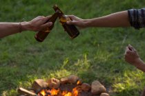 Hombre toasting beer bottle near campfire at camping - foto de stock