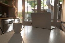 Laptop e xícara de chá na mesa na cozinha em casa — Fotografia de Stock