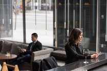 Бізнес-леді за допомогою мобільного телефону в барна стійка, а бізнесмен, використовуючи цифровий планшет на дивані в готелі — стокове фото
