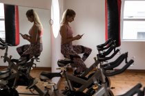 Boxer féminin utilisant un téléphone portable tout en faisant de l'exercice sur vélo d'appartement dans un studio de fitness — Photo de stock