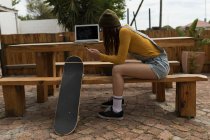 Vue latérale du skateboarder féminin utilisant le téléphone mobile au café extérieur — Photo de stock
