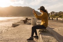Vista laterale della donna cliccando foto con cellulare vicino alla spiaggia — Foto stock