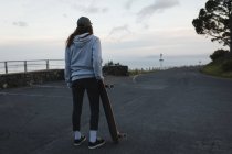 Вид сзади скейтбордиста, стоящего со скейтбордом на проселочной дороге — стоковое фото