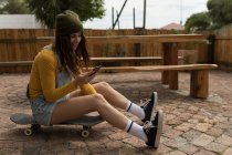 Elegante skateboarder femminile utilizzando il telefono cellulare mentre seduto sullo skateboard — Foto stock