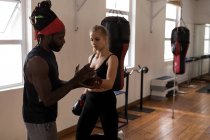 Entrenador masculino que ayuda a la boxeadora a usar guantes de boxeo en un estudio de fitness - foto de stock