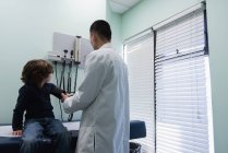 Visão traseira do jovem asiático médico masculino verificando a pressão arterial do paciente menino caucasiano na clínica. Médico masculino usando esfigmomanômetro — Fotografia de Stock