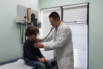 Vista lateral de un joven médico masculino asiático examinando el pecho de un niño caucásico en una clínica. Médico varón usando estetoscopio - foto de stock