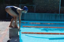 Vue latérale du jeune nageur mâle debout sur le bloc de départ en position de départ à la piscine au soleil — Photo de stock