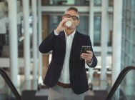 Вид спереду бізнесмена п'є каву, використовуючи свій мобільний телефон на ескалаторі в офісі — стокове фото