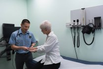 Joven médico asiático examinando a una paciente mayor en la clínica mientras se toca las manos - foto de stock