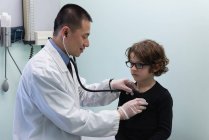 Вид сбоку на молодого врача-азиата, осматривающего белого мальчика с помощью инструмента в клинике — стоковое фото