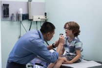Vista lateral de joven asiático médico masculino examinando caucásico niño paciente con herramienta en una clínica - foto de stock