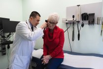 Vista frontal de un joven médico asiático examinando a un paciente mayor en la clínica - foto de stock