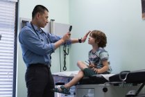Vue latérale du jeune médecin masculin asiatique examinant une oreille de patient garçon caucasien avec outil dans une clinique — Photo de stock