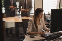 Вид збоку бізнес-леді, що працює на комп'ютері за столом в офісі з бізнесменами, що стоять на задньому плані — стокове фото