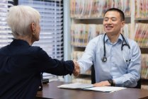 Joven asiático médico masculino y paciente senior interactuando entre sí en la clínica y estrechando la mano - foto de stock